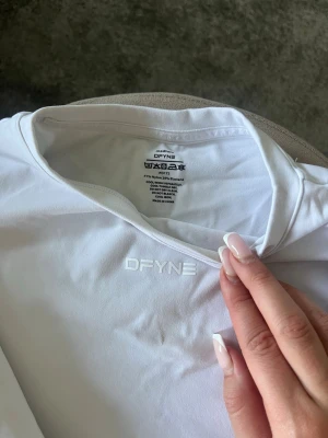 Vit långärmad topp från DFYNE - Säljer en vit långärmad topp från DFYNE med diskret logga framtill. Tillverkad i mjuk polyester och elastan för en skön och stretchig passform. Perfekt till träning eller chill dagar. Enkel och stilren design med rund hals.