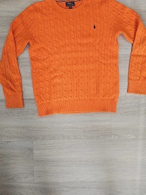 Orange kabelstickad tröja Polo Ralph Lauren - Snygg orange kabelstickad tröja från Polo Ralph Lauren med rund halsringning och klassisk marinblå logga broderad på bröstet. Tröjan har ribbade muddar vid ärmslut och nederkant. Perfekt för dig som vill sticka ut med färg i garderoben. Storlek XL men användes ett få tal gånger nör jag var 13. Pris kan diskuteras, hör gärna av er!