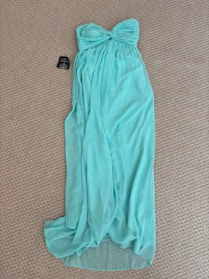 Turkos axelbandslös långklänning - Snygg turkos axelbandslös långklänning med draperad överdel och flowy kjol. Klänningen har en romantisk och luftig känsla, perfekt för speciella tillfällen. Tillverkad i ett lätt och mjukt polyestertyg som faller fint.