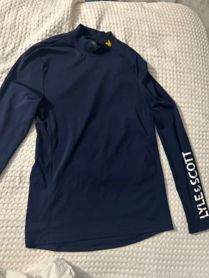 Mörkblå långärmad träningströja Lyle & Scott - Mörkblå långärmad träningströja från Lyle & Scott med vit logotyp på ena ärmen och gul broderad fågel på kragen. Tröjan har en sportig passform och är tillverkad i ett stretchigt funktionsmaterial som passar perfekt till träning.