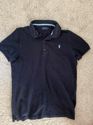 Mörkblå pikétröja från Polo Ralph Lauren - Klassisk mörkblå pikétröja från Polo Ralph Lauren med korta ärmar och den ikoniska broderade loggan på bröstet. Tröjan har krage och knappar framtill. Perfekt för en stilren och avslappnad look.
