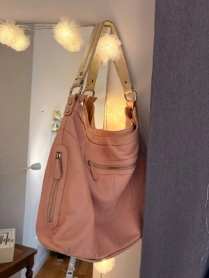 Rosa handväska med vita handtag - Säljer en ljusrosa handväska i mjukt konstläder med vita, breda handtag. Väskan har flera ytterfack med dragkedja och ett blommigt mönstrat innerfoder. Perfekt för dig som vill ha en rymlig och snygg väska till vardagen.