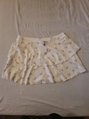 Vit blommig volangkjol - Supersöt kort kjol i vitt med små blå blommor och volangdetalj. Kjolen har en ribbad struktur och elastisk midja för skön passform. Perfekt för varma dagar och ger en romantisk vibe till din outfit.