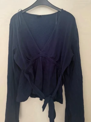 Mörkblå blus med knytband - Snygg mörkblå blus från Gina, med långa ärmar och knytband i midjan. Blusen har djup v-ringning och ett mjukt, lätt material som ger en avslappnad men chic look. Perfekt att styla med jeans eller kjol för en trendig outfit. Storlek L men passar även dig som har M. Använd fåtal gånger och är i jättebra skick. 