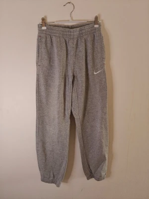 Nike mjukisbyxor  - Snygga grå mjukisbyxor från Nike med resår i midjan och vid bensluten. Byxorna har sidofickor och en vit Nike-logga broderad på ena benet. Perfekta för chill dagar eller träning. Tillverkade i mjukt och bekvämt material. Storlek M för barn.
