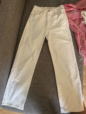 Vita raka jeans från Levi's - Säljer ett par vita raka jeans från Levi's med klassisk femficksdesign och läderpatch bak i midjan. Jeansen har hög midja och raka ben, tillverkade i kraftig bomull. Perfekta för en clean och stilren look.