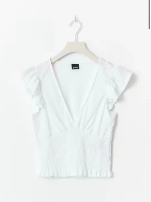 Vit croppad blus med volangärm - Superfin vit blus från Gina Tricot med v-ringning, volangärmar och smockad midja. Toppen är croppad och har en lätt rynkad kant nedtill. Perfekt för en fräsch och somrig look.