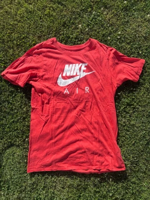 Röd Nike Air t-shirt med logga - Snygg röd t-shirt från Nike med stor vit Nike Air-logga på bröstet. Klassisk rund halsringning och korta ärmar. Perfekt för dig som gillar sportig och avslappnad stil. Tillverkad i mjuk bomull för skön känsla hela dagen.