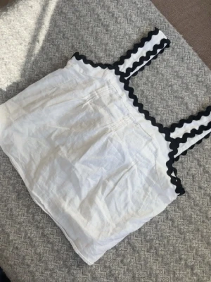 Vit topp - Söt vit linnetopp med breda axelband dekorerade med svarta vågiga detaljer. Toppen har en lös passform och knytning i ryggen för extra stil. Perfekt för varma dagar och enkel att matcha med jeans eller kjol.