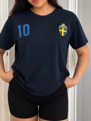 Blå Sverige t-shirt med nummer 10 - Mörkblå t-shirt med Sveriges landslagsemblem på bröstet och siffran 10 i ljusblått tryck. Klassisk rund hals och korta ärmar, perfekt för sport eller supporterhäng. Tillverkad i mjukt bomullsmaterial.