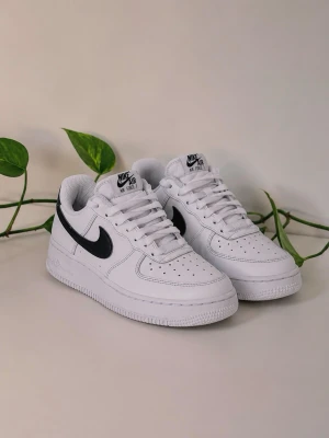 Nike Air Force 1 vita sneakers - Klassiska Nike Air Force 1 sneakers i vitt läder med svart swoosh och svart detalj på hälen. Lågt skaft, perforerad tå och vit sula ger en clean och tidlös look. Perfekta för dig som vill ha en ikonisk och stilren sneaker till vardagslooken.