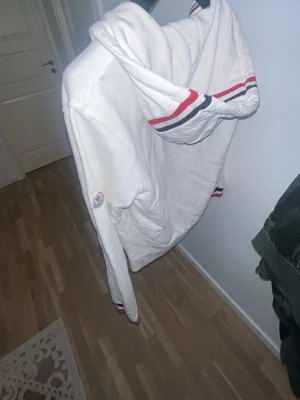 Vit hoodie från Moncler med dragkedja - Vit hoodie från Moncler med dragkedja, huva och fickor framtill. Röd, vit och marinblå randdetalj på huva, ärmslut och nederkant. Moncler-logga på ärmen. Tillverkad i mjukt bomullsmaterial, perfekt för en avslappnad och stilren look.