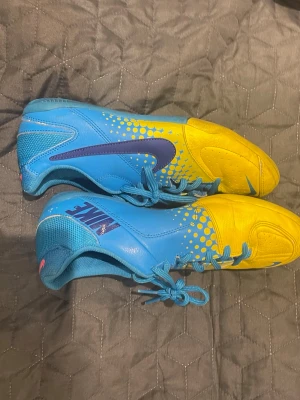 Nike blå-gula fotbollsskor - Snygga fotbollsskor från Nike i blått och gult med coola prickdetaljer och en stor mörkblå Swoosh på sidan. Skorna har snörning, slitstark yttersula med mönster för bra grepp och är tillverkade i syntetmaterial och mesh för bra ventilation.