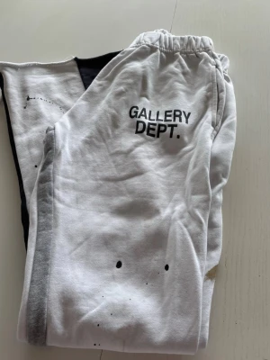Vita mjukisbyxor från Gallery Dept. - Säljer ett par vita mjukisbyxor från Gallery Dept. med svart logga på vänster sida och coola färgstänk-detaljer. Byxorna har resår i midjan och är gjorda i mjukt bomullsmaterial. Perfekta för en avslappnad och trendig look.
