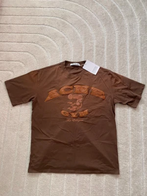 Brun Acne Studios t-shirt med tryck - Säljer en brun t-shirt från Acne Studios med coolt tryck och broderade bokstäver på bröstet. T-shirten har rund hals och korta ärmar. Perfekt för dig som gillar streetwear och vill ha något unikt i garderoben.