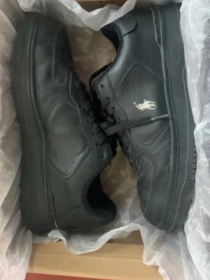 Polo Ralph Lauren Masters Court sneakers svart  - Stilrena svarta sneakers från Polo Ralph Lauren med klassisk låg siluett. Skorna är i läder med svart ovandel och har den ikoniska vita Polo-loggan på sidan. Perfekta för dig som gillar en clean och tidlös look. Köptes för några månader sedan men endast använda i någon vecka tills jag köpte ett par andra istället, dessa kommer ej till användning då de bara legat i garderoben i ett par månader nu. Nypris 1900kr
