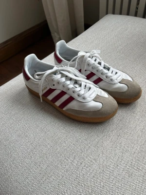 Adidas Samba vita och vinröda sneakers - Säljer ett par klassiska Adidas Samba sneakers i vitt med vinröda ränder och detaljer. Skorna har beige mocka framtill och gummisula. Ikonisk retrostil med lågt skaft och vita skosnören. 