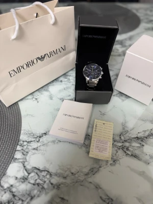 Emporio Armani silverfärgad klocka blå - Stilren klocka från Emporio Armani med blå urtavla och silverfärgat armband i metall. Klockan har kronograf-funktion och tydliga indexmarkeringar. Levereras i originalask med tillhörande manual och påse.