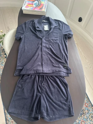 Mörkblå loungewear-set från Les Deux - Mysigt loungewear-set från Björn Borg i mörkblå frotté. Setet består av en kortärmad skjorta med knappar och bröstficka samt matchande shorts. Perfekt för chill dagar hemma eller när du vill vara bekväm men ändå snygg. Settet är helt oanvändt, därav sitter lappen fortfarande på. 