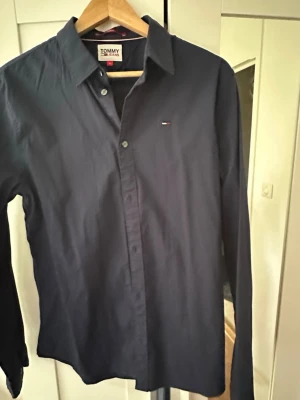 Mörkblå skjorta Tommy Jeans slim fit - Snygg mörkblå skjorta från Tommy Jeans i slim fit-modell. Klassisk design med knappar framtill och liten logga på bröstet. Perfekt till jeans eller chinos för en clean och stilren look.