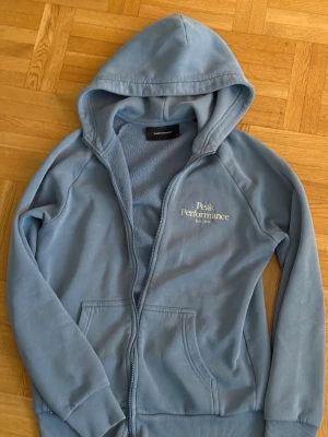 Blå hoodie från Peak Performance - Blå hoodie från Peak Performance med broderad logga på bröstet. Tröjan har huva, dragkedja och två fickor framtill. Insidan är mjuk och fleecefodrad, perfekt för kyliga dagar. Långa ärmar med ribbade muddar och klassisk passform.