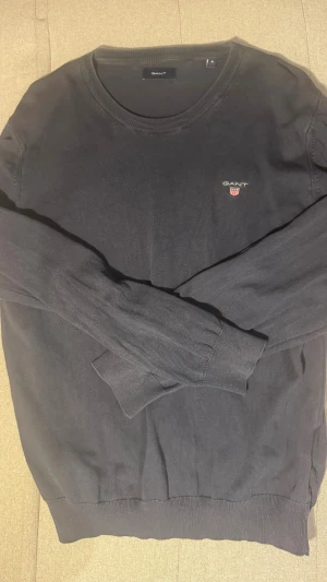 Marinblå stickad tröja från GANT - Marinblå stickad tröja från GANT i klassisk modell med rund hals och ribbade muddar. Diskret GANT-logga med broderat emblem på bröstet. Perfekt för dig som gillar stilrena och tidlösa plagg. Materialet är mjukt och skönt mot huden. Hör av dig om du har frågor!