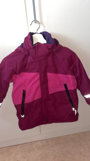 Mörklila och rosa vindjacka - Snygg vindjacka från FIX i mörklila och rosa med två dragkedjefickor och reflexdetaljer. Jackan har hög krage och mjukt fleecefoder på insidan för extra komfort. Perfekt för dig som gillar färg och vill sticka ut lite extra.