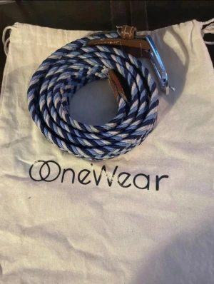 Randigt blått bälte från OneWear - Säljer ett snyggt flätat bälte från OneWear med randigt mönster i olika blå nyanser och vitt. Bältet har detaljer i brunt skinn vid spännet och en silvrig metallspänne. Perfekt för att ge din outfit lite extra edge.