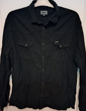 Svart Wrangler skjorta med knappar - Svart skjorta från Wrangler med klassisk krage och tryckknappar framtill. Två bröstfickor med lock och knapp, samt långärmad modell. Skjortan har en rak passform och är tillverkad i bomull, perfekt för dig som gillar enkel och stilren look.