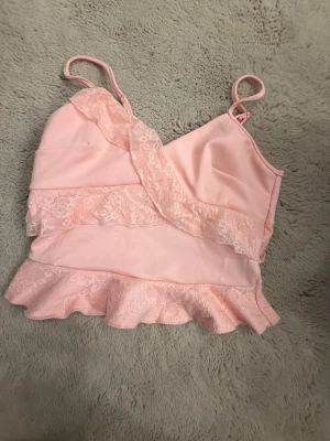 Ljusrosa linnetopp med spetsvolang - Supersöt ljusrosa linnetopp med smala axelband och omlott-design. Toppen har dekorativa spetsvolanger både framtill och längs nederkanten, vilket ger en romantisk vibe. Perfekt för dig som gillar feminina detaljer och mjuka färger.