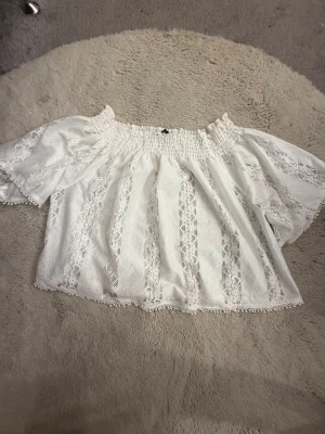 Vit offshoulder blus från Gina Tricot - Superfin vit blus från Gina Tricot i storlek XS. Offshoulder-modell med bred smock upptill och korta ärmar. Blusen har spetsdetaljer i vertikala ränder och en luftig, flowy passform. Perfekt för sommarens alla tillfällen, med romantisk vibe och snygga spetskanter.