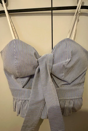 Randig korsett-topp med rosett - Säljer en blåvit randig korsett-topp med smala axelband, stor rosett framtill och volangkant nedtill. Toppen har dragkedja i ryggen och är croppad. Materialet är polyester, nylon och elastan, tillverkad i Italien. Perfekt för dig som gillar detaljer och snygg passform.