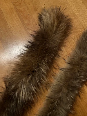 Brun fluffig pälsimitation halsduk - Säljer två riktigt fluffiga och långa pälskragen i brun pälsimitation med svarta och ljusa inslag. De har en naturlig look och är perfekta för att ge din outfit extra edge under kalla dagar. Insidan är fodrad med svart tyg och har små öglor för enkel fastsättning.