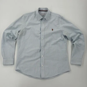 Randig skjorta från U.S. Polo - Klassisk skjorta från U.S. Polo Assn. i ljusblått och vitt randigt mönster. Skjortan har broderad logga på bröstet, knappar framtill och lång ärm med manschett. Tillverkad i mjuk bomull o har en snygg, tidlös look