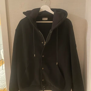 Unik moncler zip hoodie - Svart hoodie från Moncler med stor huva, snörning och både dragkedja och knappar framtill. Tröjan har en rymlig känguruficka och långärmade muddar. Materialet känns tjockt och mjukt, perfekt för kyliga dagar. Snygg och stilren design som passar till streetwear.