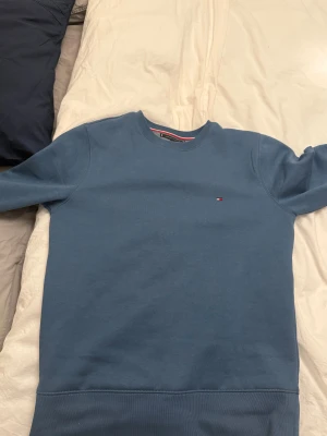 Blå sweatshirt från Tommy Hilfiger - Snygg blå sweatshirt från Tommy Hilfiger i storlek S. Tröjan har rund hals, lång ärm och ett litet broderat Hilfiger-logo på bröstet. Tillverkad i mjuk bomull med ribbade muddar vid ärmslut och nederkant. Perfekt för chill dagar samt när man vill va lite finklädd.