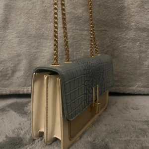 Ljusblå och beige axelväska - Snygg axelväska i blått och beige med krokopräglat lock och guldfärgad kedjerem. Väskan har två fack och ett elegant metallspänne framtill. Perfekt skick