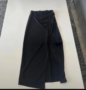 Svart långkjol med slits från Gina Tricot - Svart långkjol från Gina Tricot i slät polyester med snygg omlottkänsla och slits framtill. Kjolen har rynkade detaljer i midjan som ger en cool och modern look. Perfekt för dig som gillar stilrena och trendiga plagg.