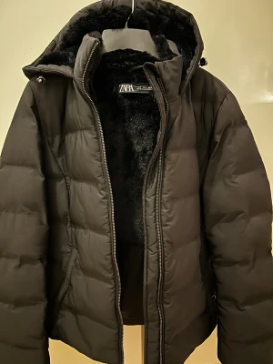 Svart pufferjacka från Zara - Säljer en svart pufferjacka från Zara med fluffigt foder och huva. Jackan har dragkedja framtill och en stilren, puffig design som håller dig varm. 