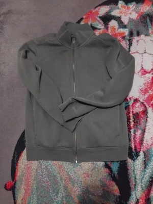 Grå Sail Racing ziptröja - Grå ziptröja från Sail Racing med hög krage och broderad logga på bröstet. Tröjan har hel dragkedja framtill och är tillverkad i en mjuk blandning av bomull och polyester. Perfekt för dig som gillar stilren och enkel design.