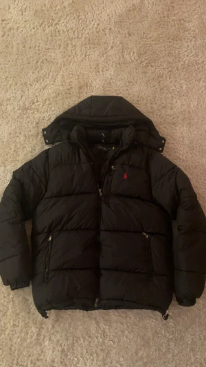 Svart pufferjacka från Polo Ralph Lauren - Svart pufferjacka från Polo Ralph Lauren med röd broderad logga på bröstet. Jackan har huva, flera dragkedjeförsedda fickor och quiltad design. Perfekt för kalla dagar med sin tjocka och fluffiga look. Klassisk stil med snygga detaljer och långärmad modell.  PRISET KAN DISKUTERAS! Kom gärna med frågar!:)