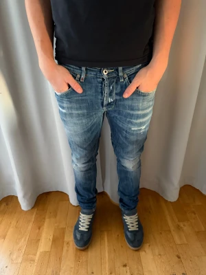 Blåa dondups med slitningar. - Säljer ett par blå skinny jeans från George i storlek 33. Jeansen har slitna detaljer och tvättad look, klassisk femficksmodell och knappgylf. Snygg passform som sitter tajt längs benen och ger en modern vibe. Perfekt för dig som gillar en smal siluett.