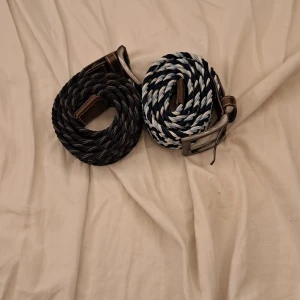 Flätade bälten med spänne - Två stiliga flätade bälten med metallspänne. Ett bälte är svart och det andra har ett mönster i blått och vitt. Perfekta för att ge en extra touch till din outfit.