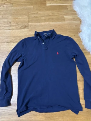 Marinblå långärmad piké från Polo Ralph Lauren - Snygg marinblå långärmad pikétröja från Polo Ralph Lauren i slim fit. Tröjan har klassisk krage, två knappar och den ikoniska röda logon broderad på bröstet. Tillverkad i 100% bomull för skön känsla och stilren look.