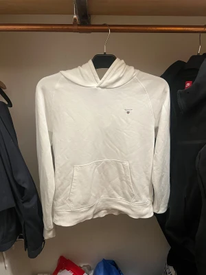 Vit hoodie från GANT med huva - Vit hoodie från GANT med klassisk huva och magficka. Tröjan har ett litet broderat GANT-logo på bröstet och är tillverkad i mjuk bomull. Långärmad och med ribbade muddar vid ärmslut och nederkant. Perfekt för chill dagar och enkel att matcha.