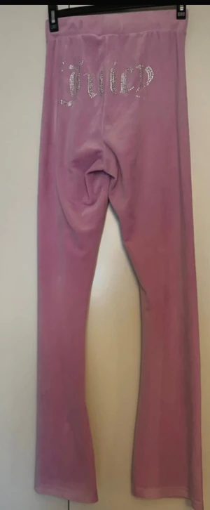 Rosa velourbyxor Juicy Couture - Säljer ett par ikoniska rosa velourbyxor från Juicy Couture med strassdetaljer på bakfickan och ett litet J framtill. Byxorna har bootcut-snitt och bred resår i midjan för extra komfort. Perfekta för en chill och trendig look.