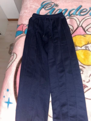Mörkblå mjukisbyxor med snörning - Säljer ett par mörkblå mjukisbyxor med raka ben och bekväm resårmidja med snörning. Byxorna har sydda vertikala sömmar längs benen som ger en cool detalj. Perfekta för chill dagar eller när du vill vara extra comfy.