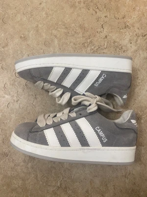 Grå Adidas Campus sneakers - Säljer ett par grå Adidas Campus sneakers med klassiska vita ränder på sidan och vit Adidas-logga på hälen. Skorna har platt sula, rund tå och är gjorda i mocka med snörning. Perfekt för dig som gillar retrostil och vill ha bekväma sneakers med snygg design.