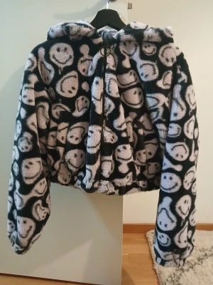 Smiley x H&M teddyjacka med huva - Fluffig teddyjacka från Smiley x H&M i svart och ljusbeige med smiley-mönster över hela jackan. Jackan har huva, dragkedja framtill och är croppad i modellen. Perfekt för dig som gillar statement-plagg och mjuka material.
