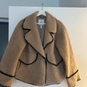 Beige teddyjacka från Object stl 36 - !!!Kan gå ner i pris!!!!Supermjuk och trendig teddyjacka från Object i beige med svarta detaljer längs kanterna. Jackan har bred krage, markerade sömmar och två fickor framtill. Perfekt för att hålla sig varm och snygg under kyliga dagar.